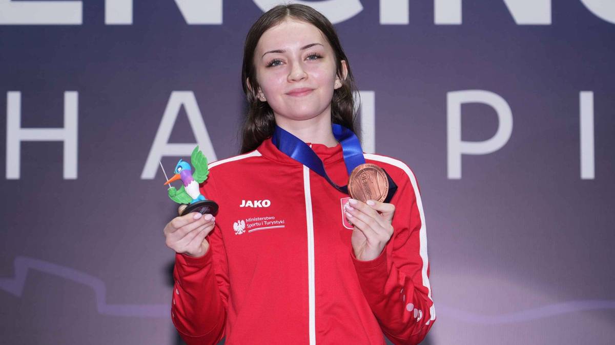 Uśmiechnięta młoda kobieta w stroju szermierczym trzymająca brązowy medal i kolorową figurkę.
