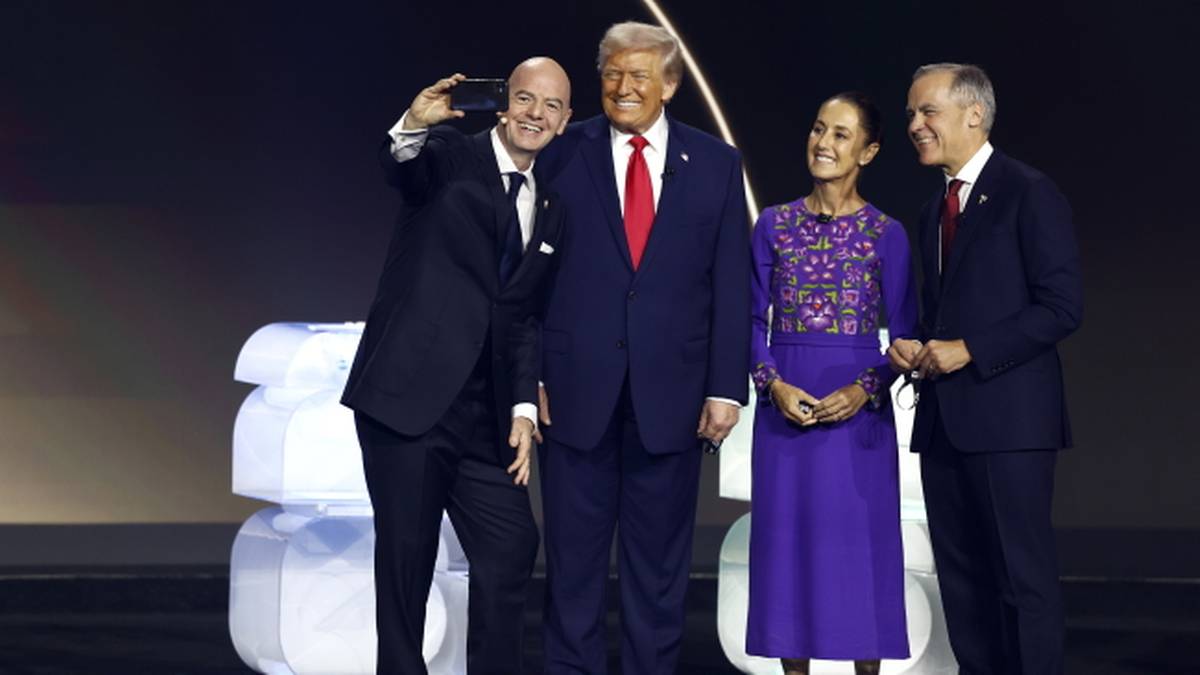 Czterech mężczyzn i jedna kobieta pozują do zdjęcia na scenie. W środku stoi Donald Trump, ubrany w granatowy garnitur i czerwony krawat. Obok niego po prawej stoi kobieta w fioletowej sukience. Po lewej stronie Trumpa stoi mężczyzna z telefonem, robiący selfie. Obok niego stoi kolejny mężczyzna.