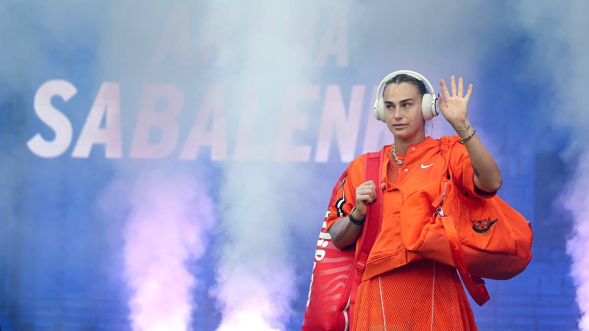 Aryna Sabalenka w pomarańczowym stroju sportowym, z torbą i słuchawkami, wchodzi na kort tenisowy.