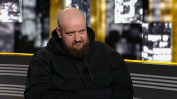 Mężczyzna z łysiną i brodą w czarnej bluzie z kapturem uśmiecha się krzywo.