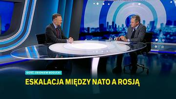 Dwaj mężczyźni w garniturach siedzą naprzeciwko siebie przy owalnym stole w studiu telewizyjnym na tle niebieskiej scenografii z grafiką miasta.
