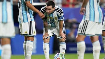 Piłkarz Lionel Messi w białej koszulce z niebieskimi pasami, pochylony nad piłką na boisku piłkarskim.