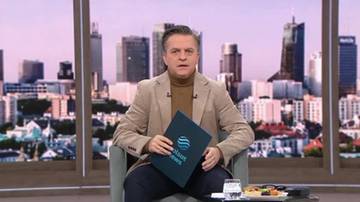 Mężczyzna w beżowej marynarce i golfie siedzi na krześle, trzymając w rękach ciemnogranatową teczkę z logo "Puls Biznesu". W tle widać panoramę miasta z wieżowcami.