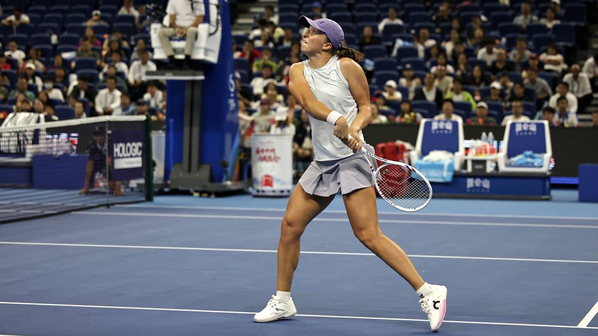 Tenisistka Iga Świątek podczas meczu, w białej bluzce i szarej spódniczce, z rakietką w dłoni, na korcie tenisowym.