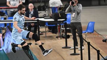 Siatkarz w niebieskim stroju wykonuje atak podczas meczu siatkówki, w tle widać fotoreportera z aparatem.