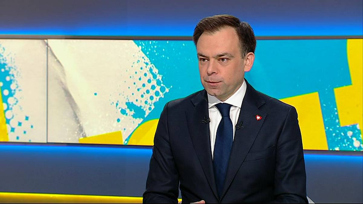 Mężczyzna w garniturze i krawacie, Andrzej Domański, siedzi przed ekranem z kolorowym wzorem.