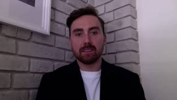 Mężczyzna z zarostem, w czarnym garniturze i białej koszulce, stoi na tle ściany z cegły.