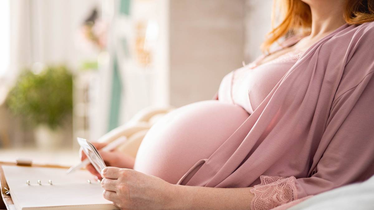 Kobieta w ciąży siedzi, trzymając w dłoniach mały przedmiot, na pierwszym planie widoczny zaokrąglony brzuch.