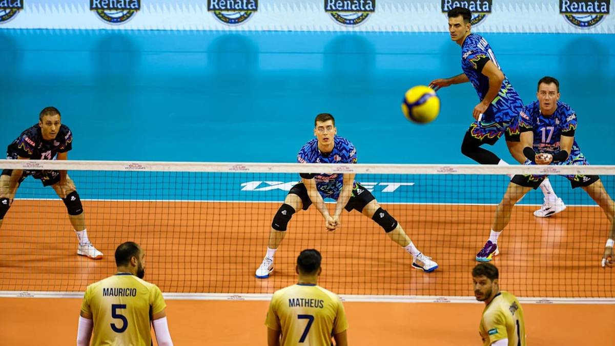 Siatkarze podczas meczu, jeden z zawodników zagrywa piłkę nad siatką, podczas gdy inni gracze przygotowują się do obrony.