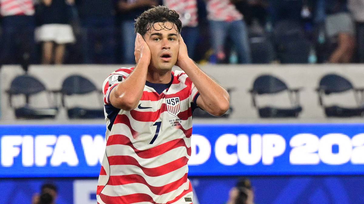 Piłkarz reprezentacji USA w czerwono-białym stroju z numerem 7 na plecach, z dłońmi przy uszach, na tle trybun z kibicami i baneru "FIFA WORLD CUP 2023".