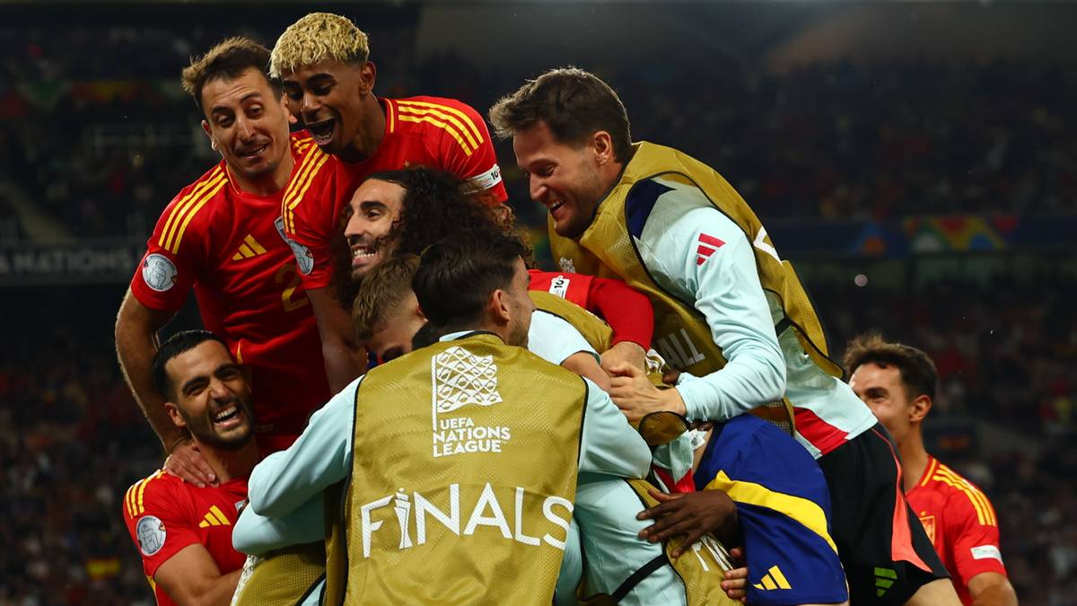 Grupa piłkarzy świętuje zwycięstwo na boisku, niektórzy trzymają się za ramiona i są na sobie, jeden z nich nosi kamizelkę z napisem 'FINALS'.