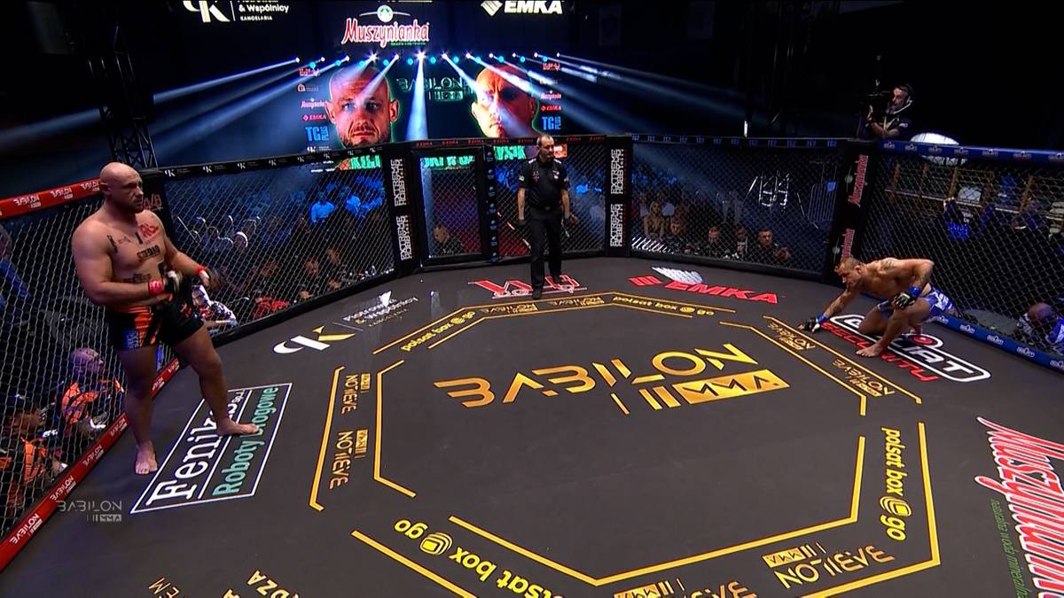 Dwóch zawodników MMA na ringu w trakcie walki na gali Babilon MMA. Jeden z zawodników klęczy na macie, drugi stoi.