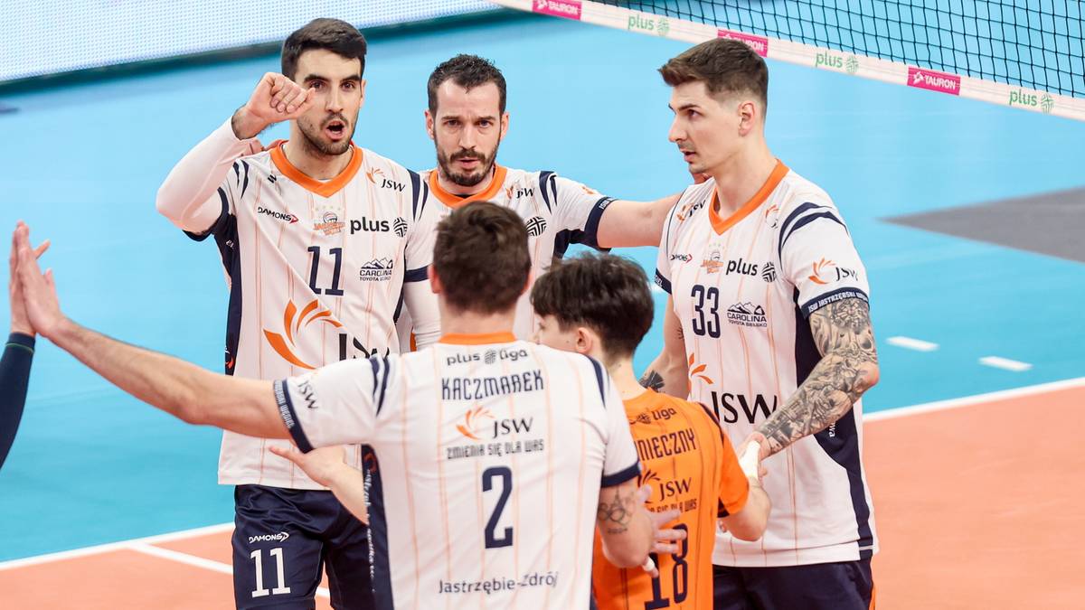 Siatkarze Jastrzębskiego Węgla w biało-pomarańczowych strojach rozmawiają ze sobą na boisku, w tle widoczna siatka.