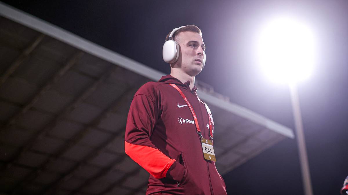 Młody mężczyzna w słuchawkach na uszach, ubrany w sportową bluzę, stoi pod jasnym światłem.