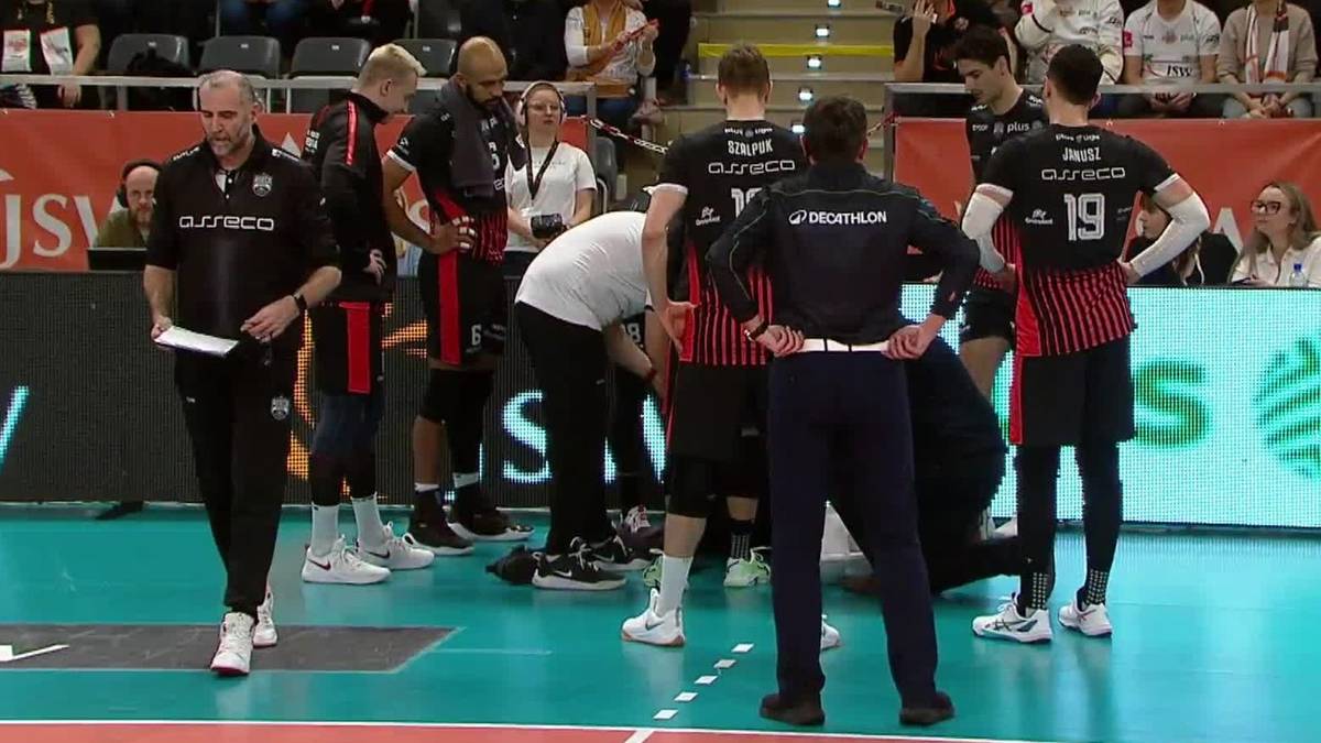 Grupa siatkarzy w czarnych strojach z czerwonymi elementami, otoczona sztabem trenerskim, podczas przerwy w meczu.