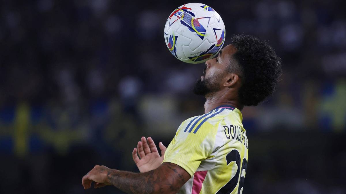 Piłkarz z kr&oacute;tkimi czarnymi włosami i brodą w ż&oacute;łtej koszulce, odbija głową białą piłkę z kolorowymi wzorami.