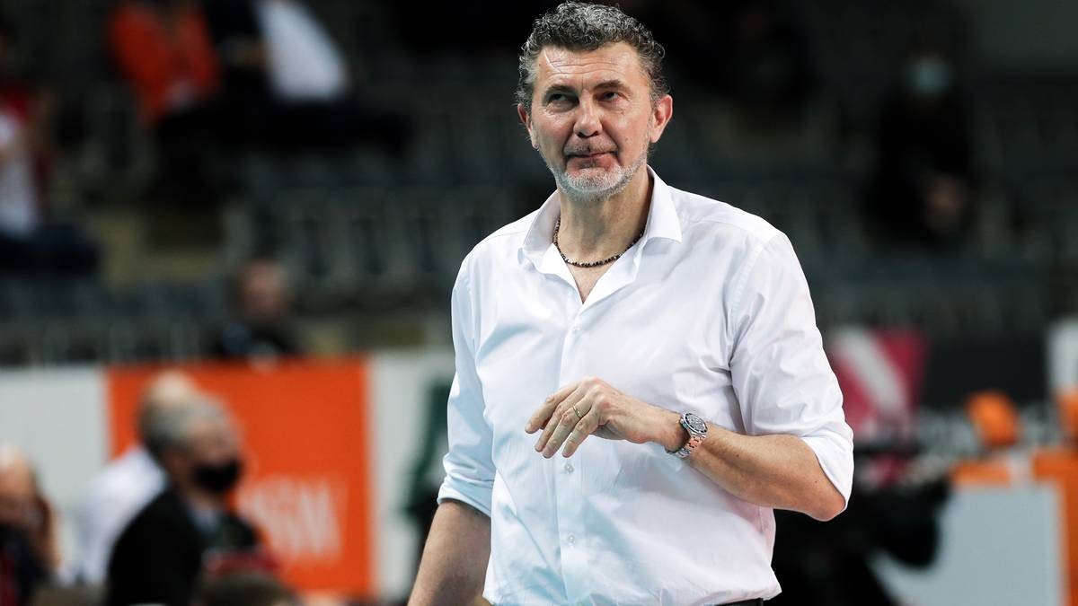 Mężczyzna w białej koszuli i ciemnych spodniach, patrzący w bok, stoi na boisku siatkarskim.