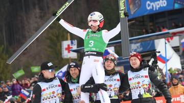 Kamil Stoch w zielonym plastronie z numerem 3, otoczony przez innych skoczk&oacute;w i trener&oacute;w, trzymający narty na tle tłumu kibic&oacute;w.