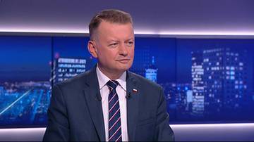 Mężczyzna w garniturze i krawacie z polską flagą przypiętą do klapy marynarki.