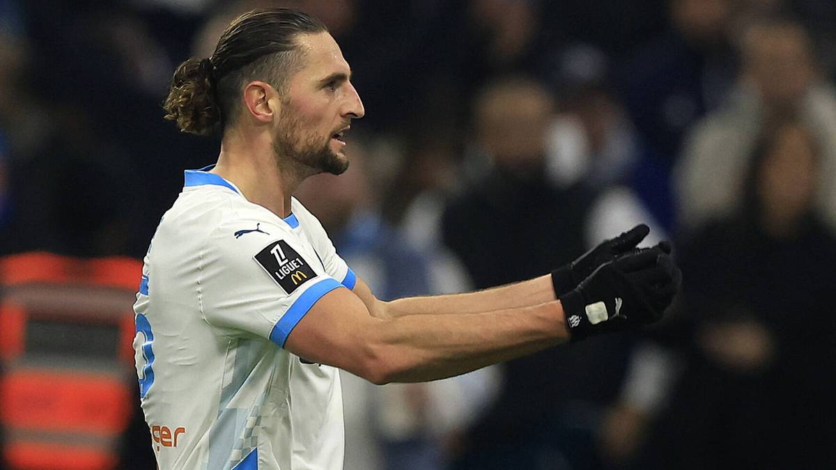 Piłkarz w białej koszulce z niebieskimi elementami i rękawiczkami, z włosami związanymi w kok, spogląda w bok z zaciętą miną.