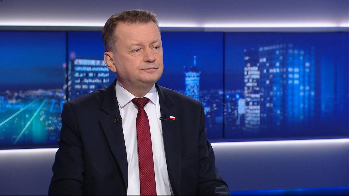 Mężczyzna w ciemnym garniturze i czerwonym krawacie siedzi na tle nocnego miasta z widokiem na drapacze chmur.