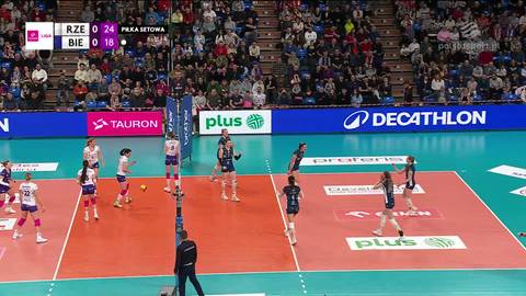 Obraz przedstawia mecz siatk&oacute;wki kobiet, w kt&oacute;rym drużyna BKS Bostik ZGO Bielsko-Biała gra przeciwko Developres Rzesz&oacute;w. Widoczne są zawodniczki na boisku, publiczność na trybunach oraz reklamy sponsor&oacute;w.