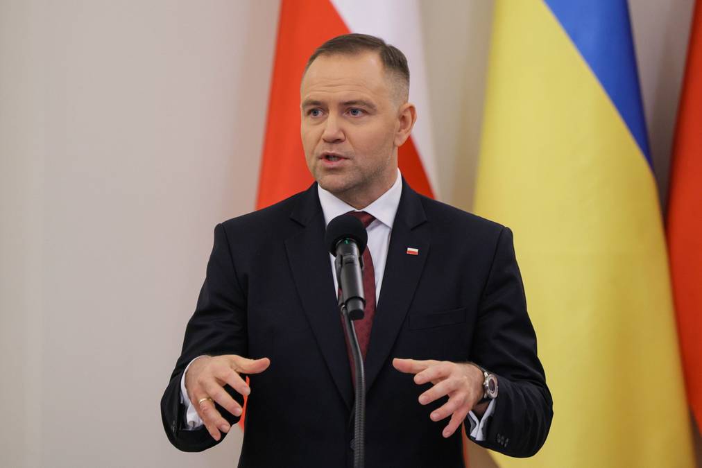 Mężczyzna w garniturze przemawia przy mikrofonie, za nim widoczne są flagi Polski i Ukrainy.