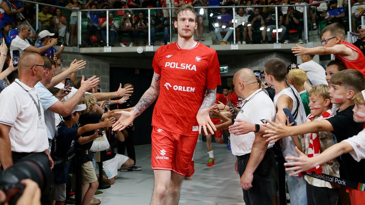 Zawodnik koszykówki w czerwonej koszulce reprezentacji Polski "POLSKA" i "ORLEN" wchodzi w otoczeniu kibiców, machając rękami.