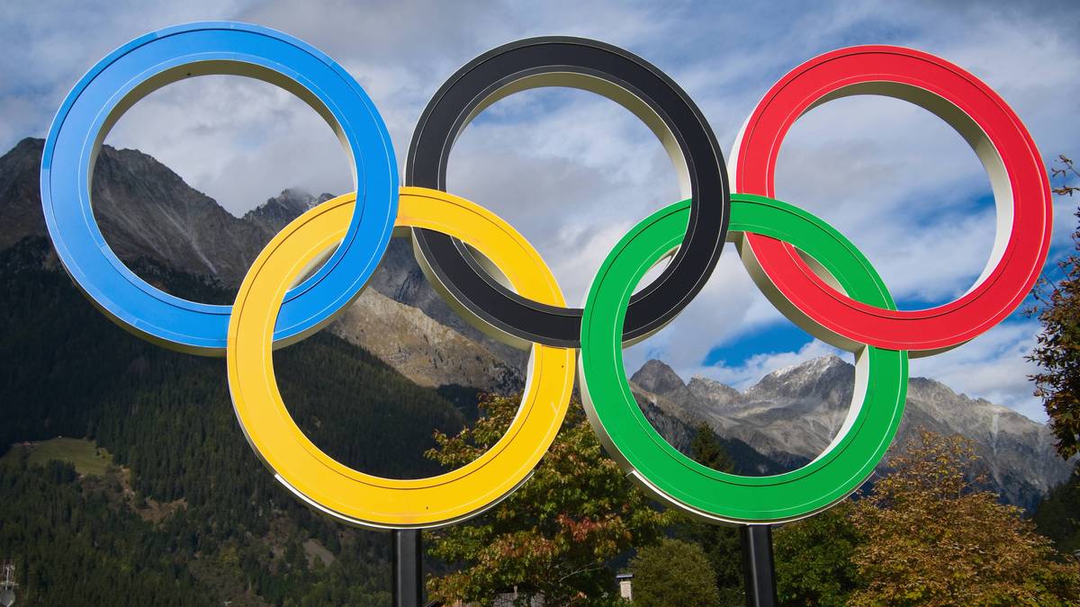 Pięć złączonych k&oacute;ł olimpijskich w kolorach niebieskim, ż&oacute;łtym, czarnym, zielonym i czerwonym, na tle g&oacute;rskiego krajobrazu z drzewami.