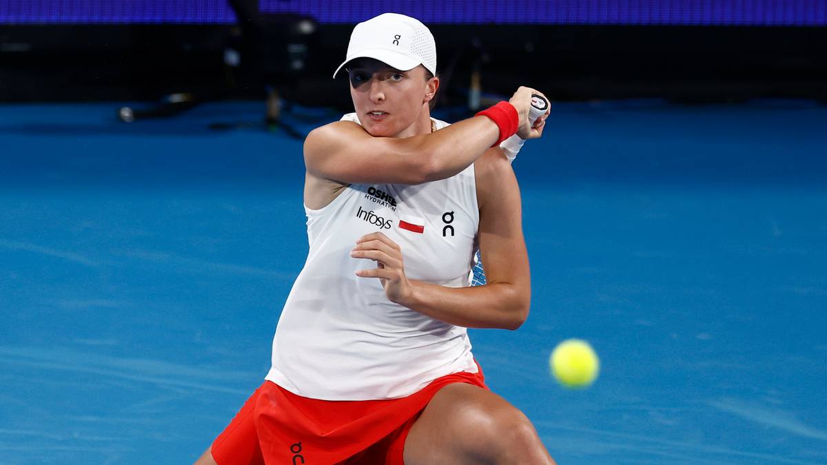 Tenisistka Iga Świątek w białej koszulce i czerwonej sp&oacute;dniczce uderza piłkę rakietą na niebieskim korcie tenisowym.