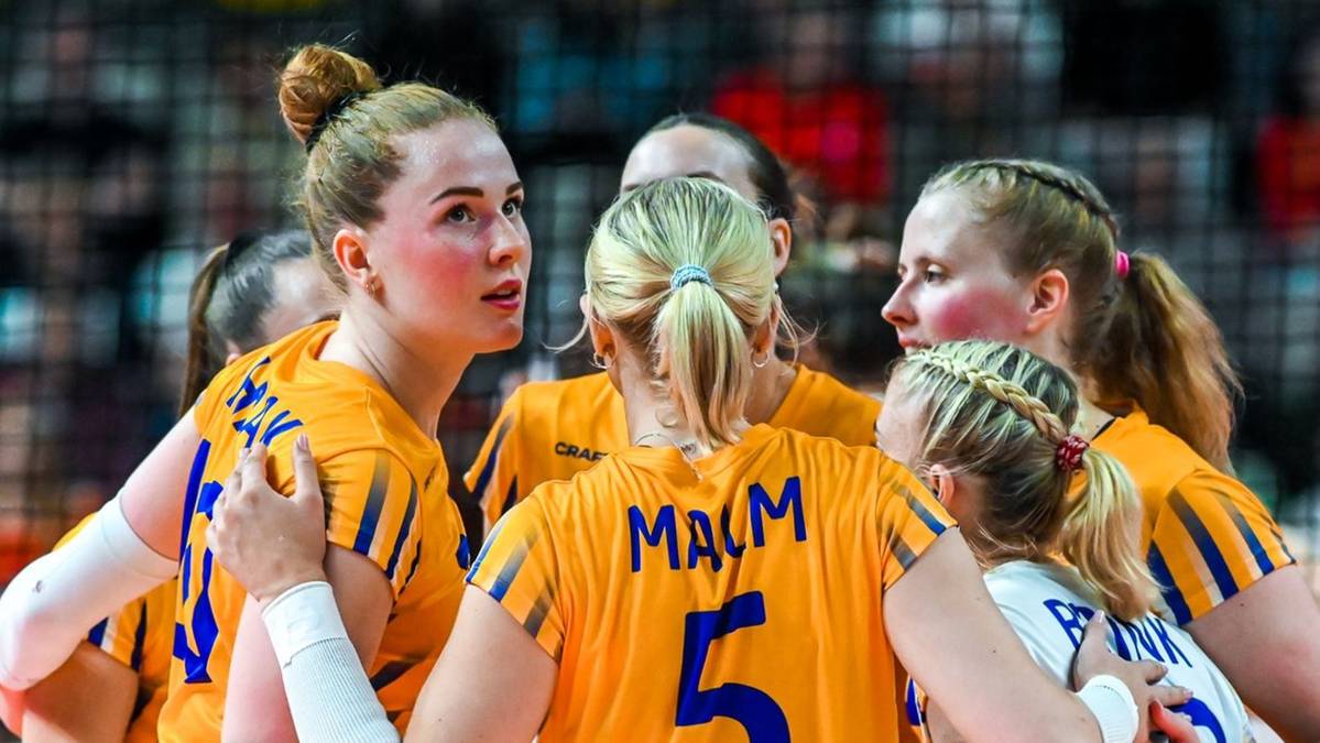 Grupa siatkarek w pomarańczowych koszulkach z białymi numerami na plecach, patrzą na siebie w skupieniu podczas przerwy w meczu.