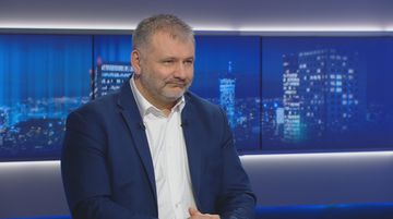 Mężczyzna w średnim wieku w białej koszuli i granatowej marynarce siedzi na tle ekran&oacute;w wyświetlających nocną panoramę miasta.