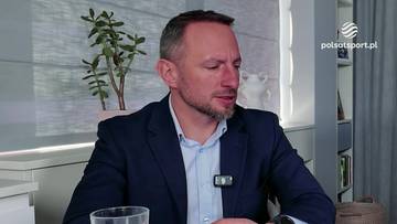 Mężczyzna w ciemnym garniturze i jasnej koszuli z mikrofonem przypiętym do kołnierzyka, siedzi za stołem z kubkiem.