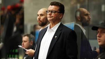 Mężczyzna w okularach i garniturze stoi w pobliżu innych mężczyzn, patrząc w dal.