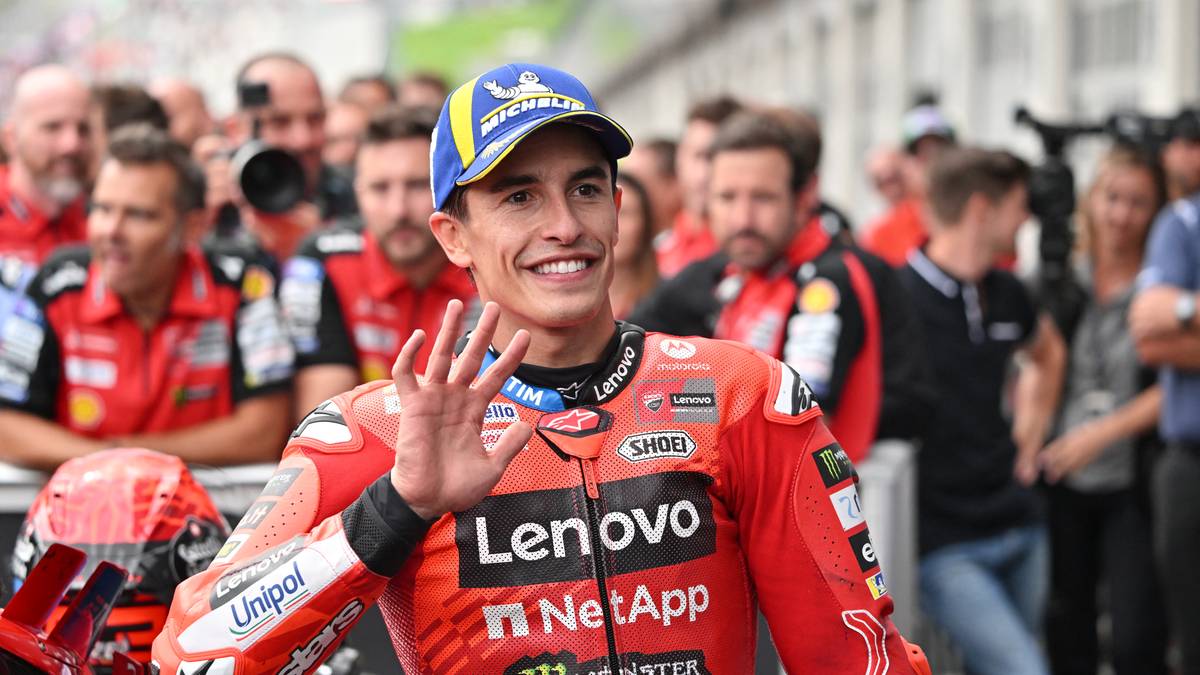 Marc Marquez, motocyklista MotoGP, w czerwono-białym kombinezonie wyścigowym i niebieskiej czapce z daszkiem, uśmiecha się i macha prawą ręką do tłumu ludzi w tle.