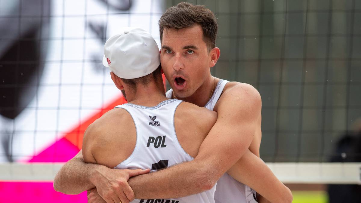 Dw&oacute;ch mężczyzn w strojach do siatk&oacute;wki plażowej przytulających się, z zawodnikiem po prawej z szeroko otwartymi ustami.