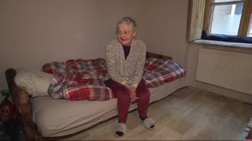 Starsza kobieta siedzi na ł&oacute;żku w pokoju, ubrana w sweter i legginsy.
