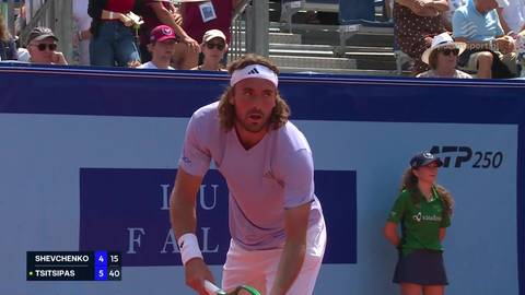 Stefanos Tsitsipas podczas meczu tenisowego, przygotowujący się do serwisu. W tle widoczne są reklamy ATP Tour 250 oraz trybuny z widzami.