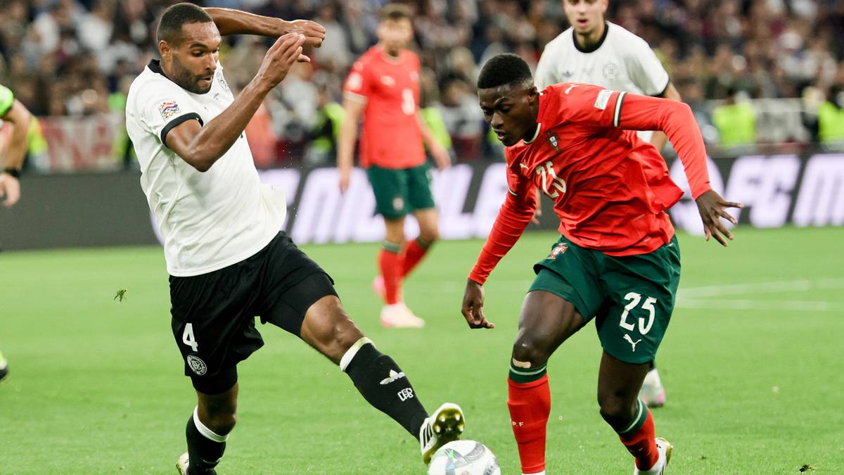 Dwóch piłkarzy walczących o piłkę na boisku piłkarskim. Jeden zawodnik w białej koszulce i czarnych spodenkach próbuje odebrać piłkę drugiemu, który ma na sobie czerwoną koszulkę i zielone spodenki z numerem 25.