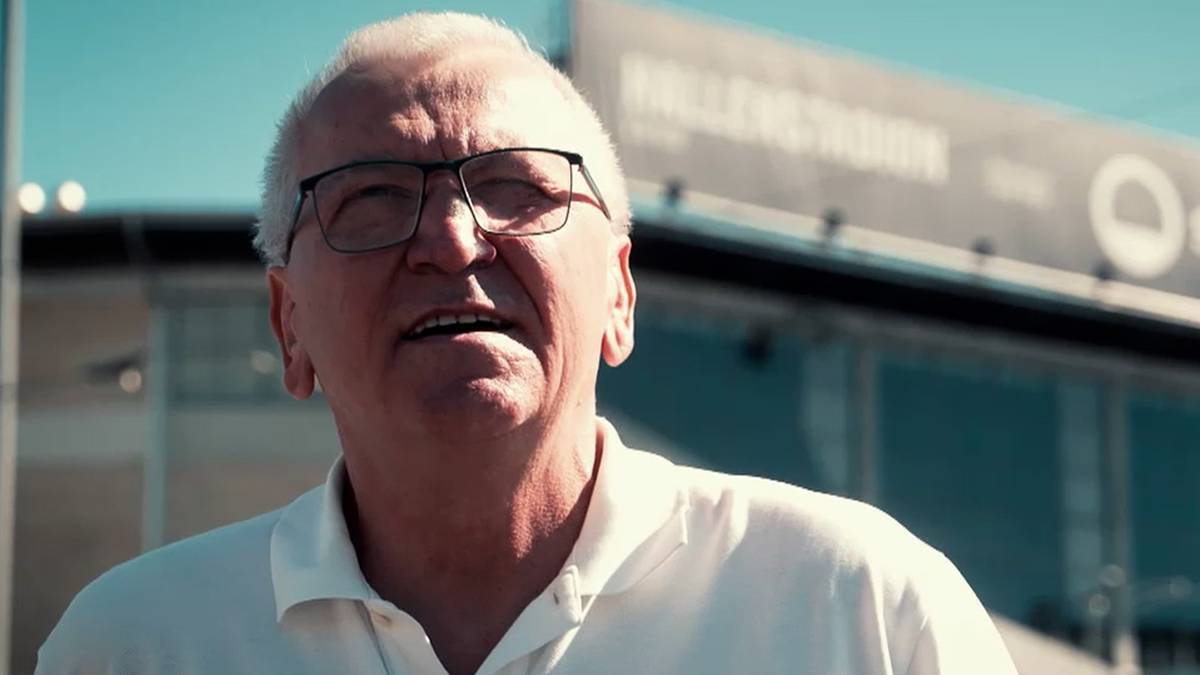 Portret starszego mężczyzny z siwymi włosami i okularami, m&oacute;wiącego z ekspresją. W tle widać fragment stadionu.