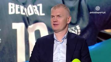 Mężczyzna w garniturze udziela wywiadu na tle drużyny siatkarskiej.