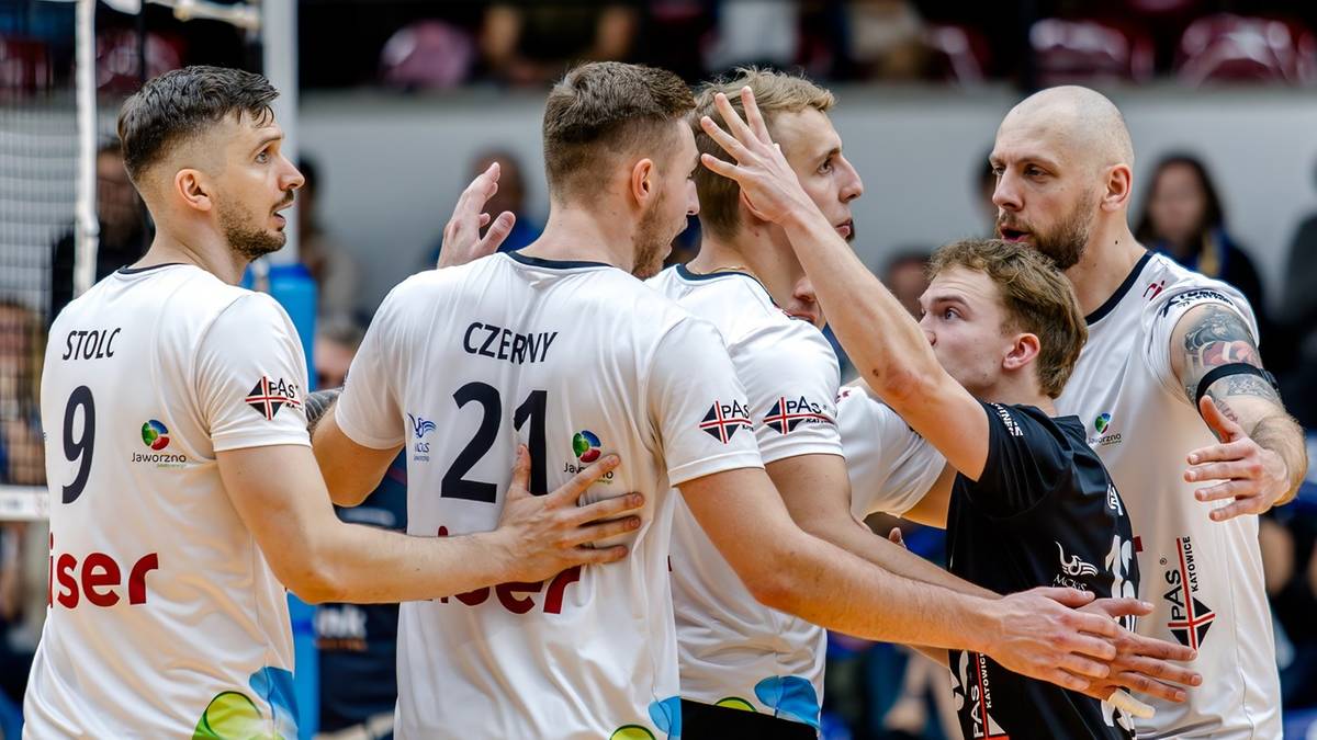 Grupa siatkarzy w białych koszulkach podczas meczu, jeden z nich podnosi rękę, inni stoją wok&oacute;ł, w tle trybuny z kibicami.