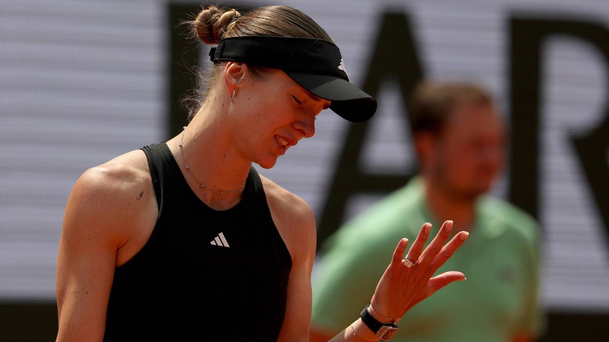 Tenisistka w czarnym stroju sportowym z opaską na głowie pochyla głowę i wyciąga dłoń w geście zamyślenia lub frustracji.