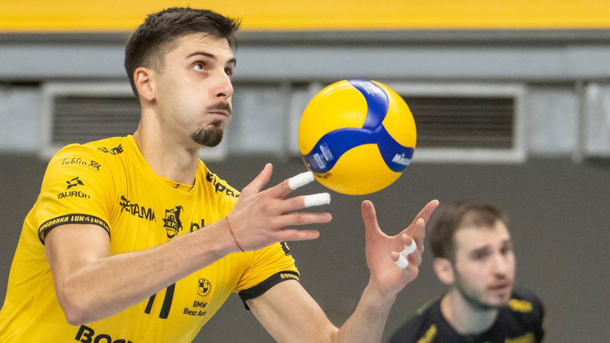 Siatkarz w żółtej koszulce z numerem 11 na piersi, przygotowuje się do zagrywki lub przyjęcia piłki do siatkówki. Piłka unosi się przed nim.