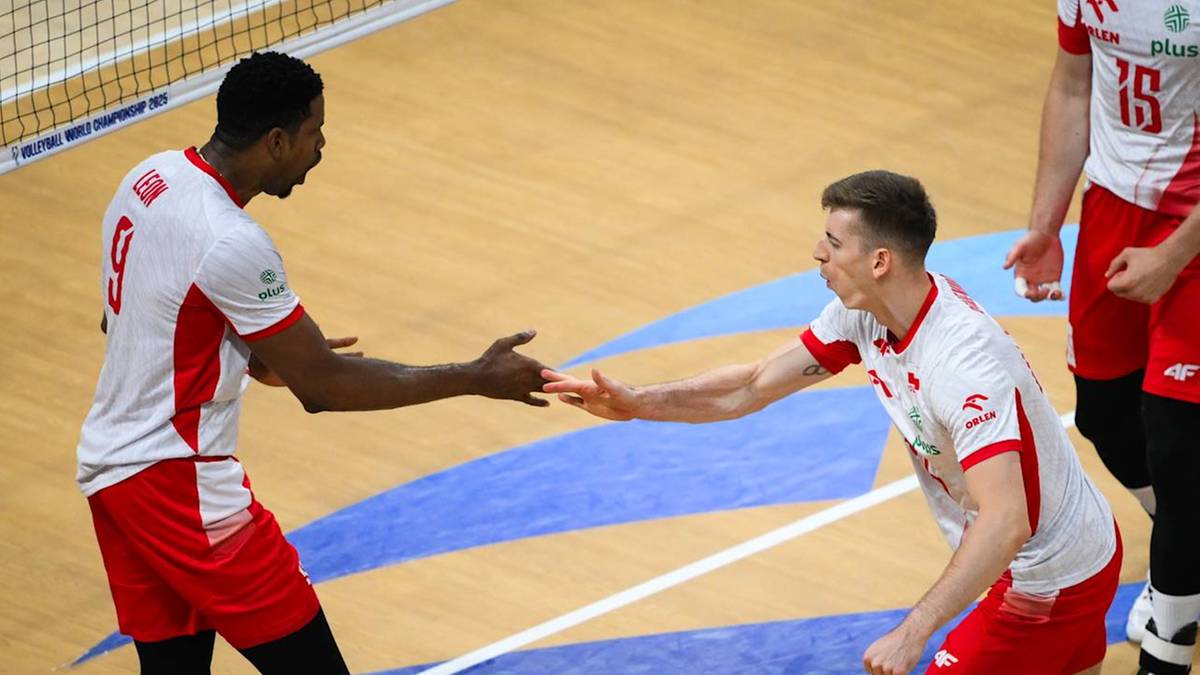 Dwóch siatkarzy w białych koszulkach z czerwonymi detalami i numerami na plecach, z dłońmi wyciągniętymi do powitania, na parkiecie hali sportowej.