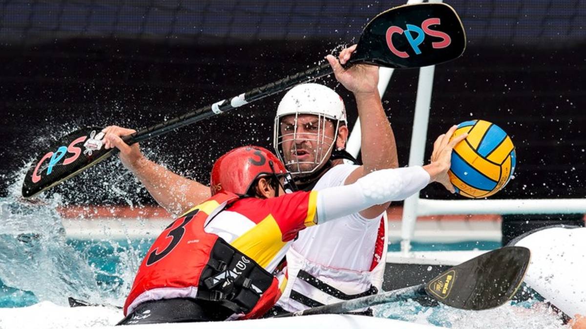 Dwóch zawodników kajak-polo walczących o piłkę w wodzie. Jeden zawodnik w białym kasku próbuje odebrać piłkę, drugi zawodnik w czerwonym kasku broni się kajakiem.