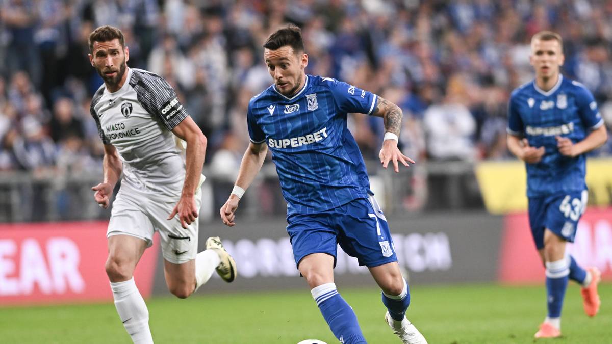 Trzech piłkarzy grających w piłkę nożną na boisku. Dw&oacute;ch w niebieskich strojach, jeden w szarym.