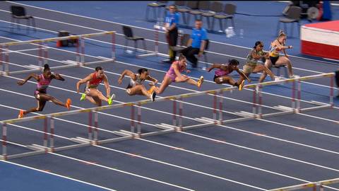 Sześć biegaczek pokonuje płotki na niebieskiej bieżni lekkoatletycznej.