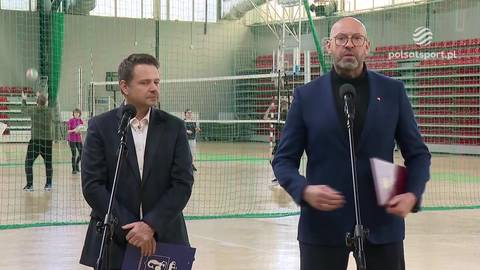 Dwaj mężczyźni w ciemnych garniturach stoją przed mikrofonami na tle hali sportowej. W tle widoczne są boisko do siatk&oacute;wki z siatką oraz grające osoby.