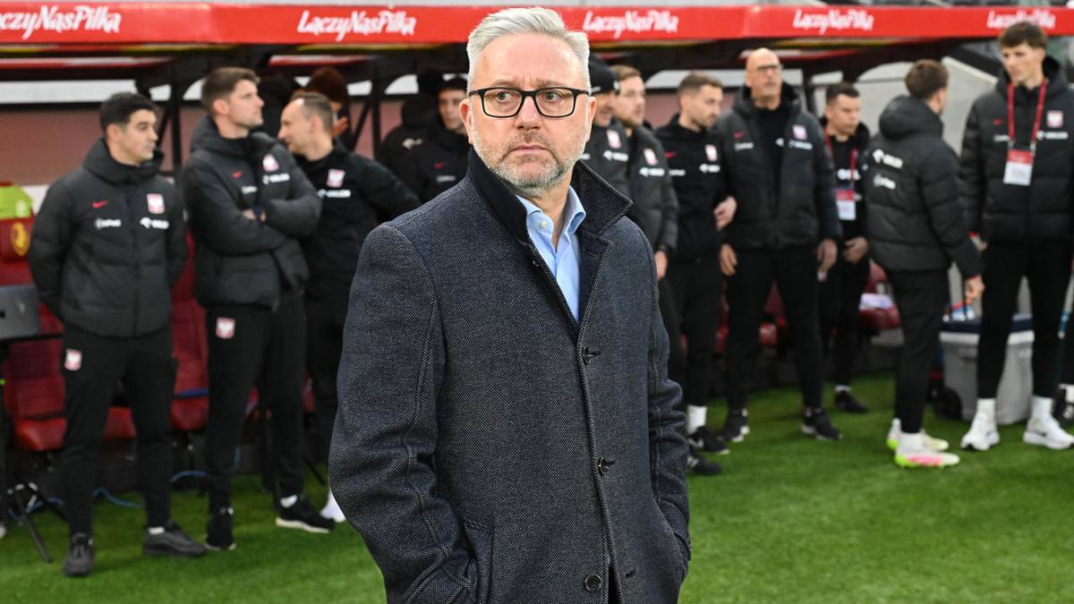 Mężczyzna w okularach i marynarce stoi na boisku piłkarskim, za nim grupa piłkarzy w dresach.